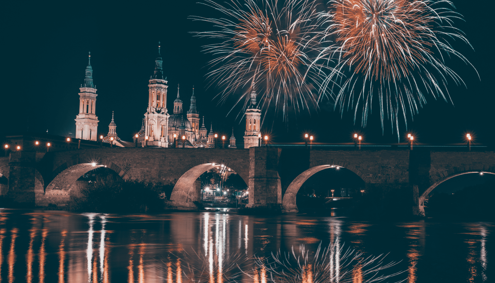 El Puente de Piedra y la Basílica de Nuestra Señora del Pilar de Zaragoza iluminados por fuegos artificiales que se reflejan en el río Ebro de noche