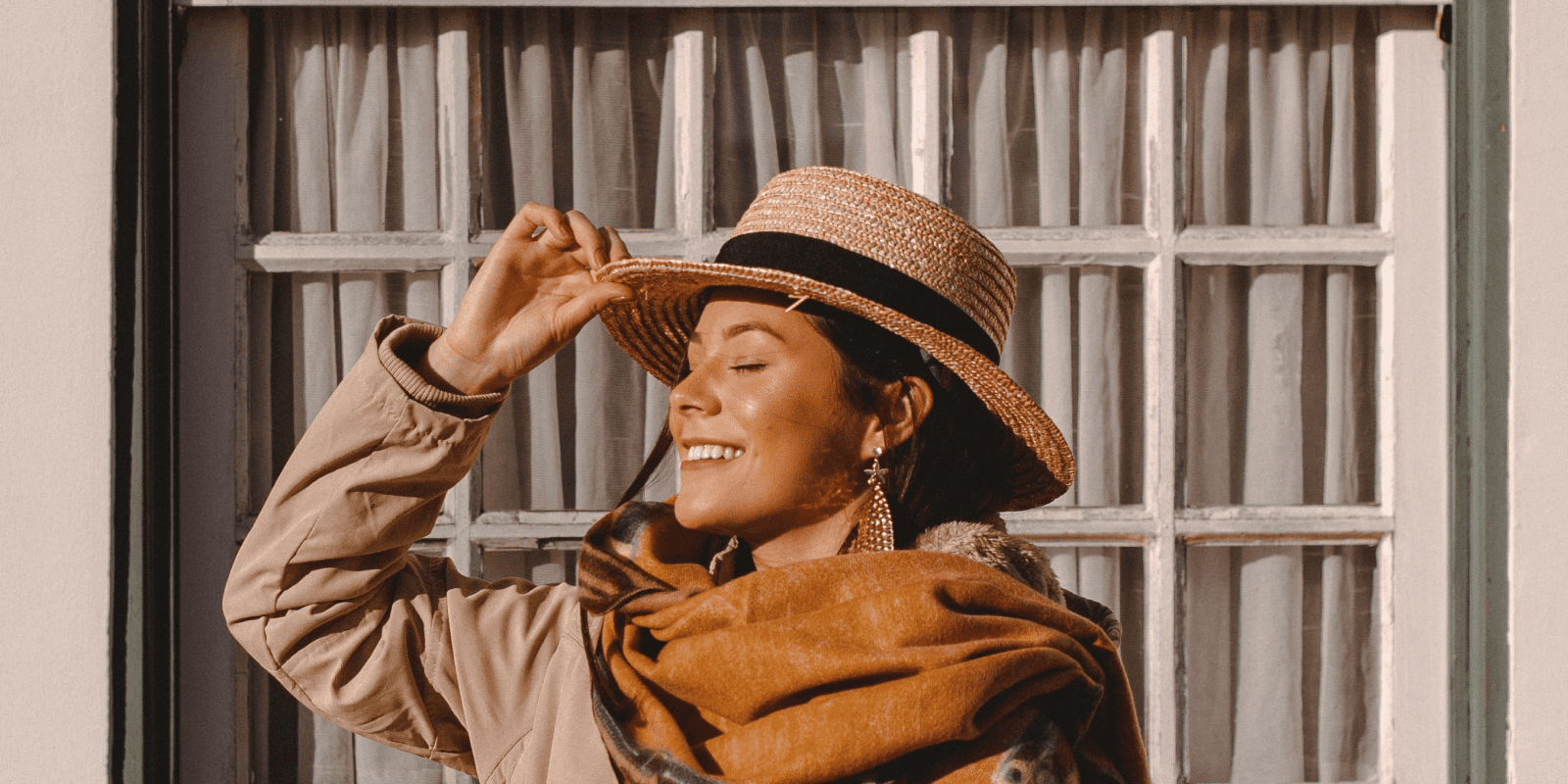 Mujer sonriente con sombrero toma el sol frente a una ventana.