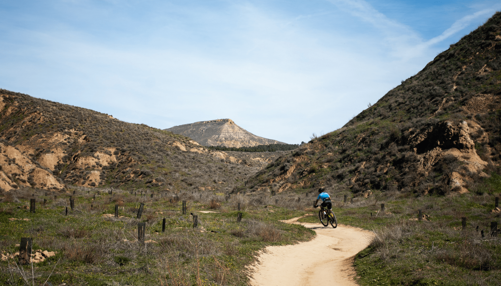 Persona en bicicleta avanza por un camino terroso rodeado de naturaleza silvestre y montañas.