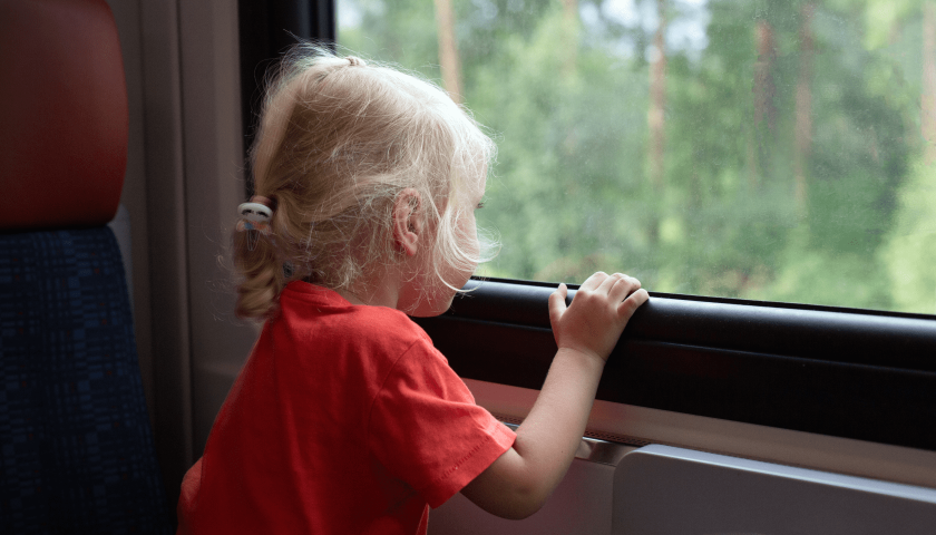 Niña pequeña rubia mira por la ventana un paisaje con árboles.