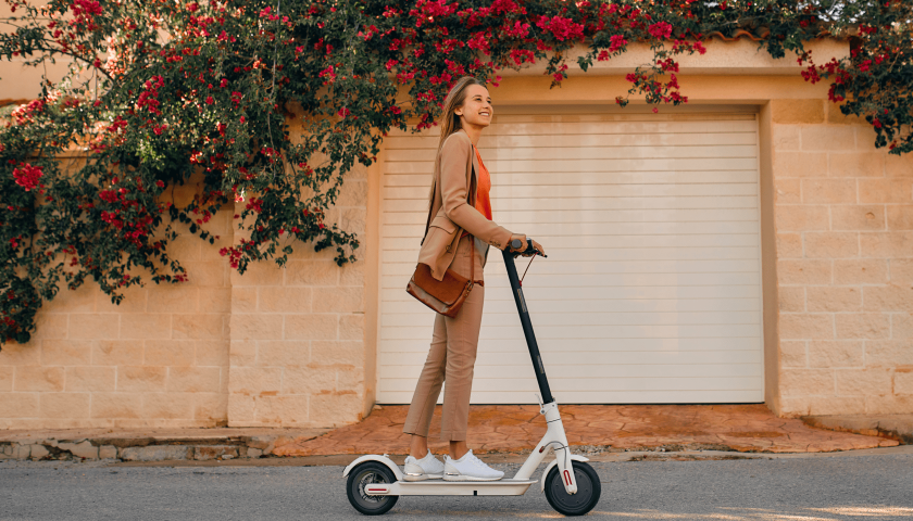 Joven vestida de marrón disfruta de un viaje en patinete eléctrico por una calle que tiene flores rojas en la fachada.